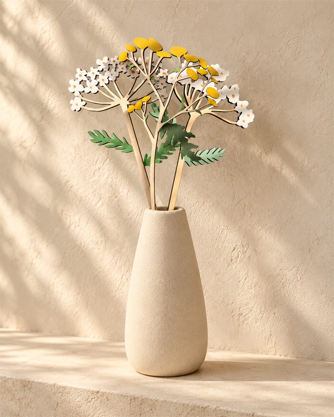 White wooden flowers, golden stems and green leaves arranged in a pale vase against a warm plaster wall